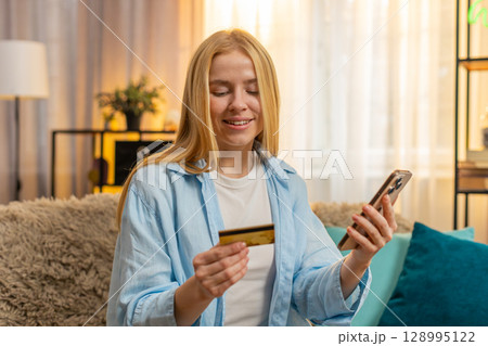 Young woman paying with credit card, smartphone transaction, relief, success at home couch, sunset Young woman paying with credit card, smartphone transaction, relief, success at home couch, sunset 128995122