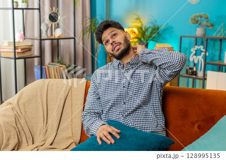 Tired ill sick Indian young man suffers from spine neck muscle discomfort pain at home room sofa 128995135