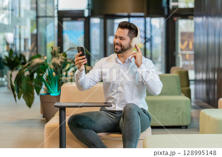 Excited young businessman in headphones listening energetic music on smartphone in office lobby 128995148