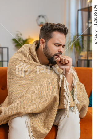 Sick Caucasian young man covered with blanket coughing while sitting on sofa in living room Sick Caucasian young man covered with blanket coughing while sitting on sofa in living room 128995206