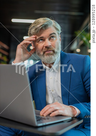 Bored sleepy mature Caucasian businessman working on laptop computer sitting in modern office lobby 128995218