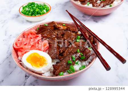 Gyudon Japanese beef with rice, pickled ginger, boiled egg slice, garnished with green onions, in a bowl with chopsticks, Gyudon Japanese beef with rice, pickled ginger, boiled egg slice, garnished with green onions, in a bowl with chopsticks, 128995344