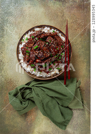 crispy beef, with green onions and sesame seeds, on a rice pad, in a bowl with chopsticks, on a textured surface, no people crispy beef, with green onions and sesame seeds, on a rice pad, in a bowl with chopsticks, on a textured surface, no people 128995345