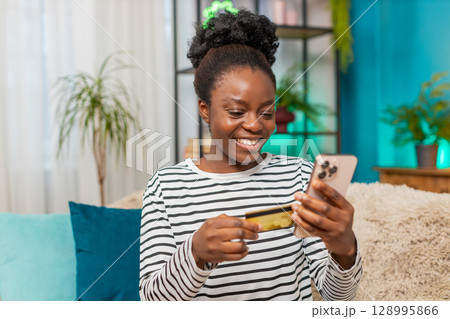 African American woman shopping online smiling entering credit card details on smartphone at home African American woman shopping online smiling entering credit card details on smartphone at home 128995866