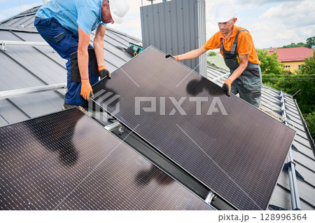Workers building solar panel system on rooftop of house. Two men installers in helmets installing photovoltaic solar module outdoors. Alternative, green and renewable energy generation concept. 128996364