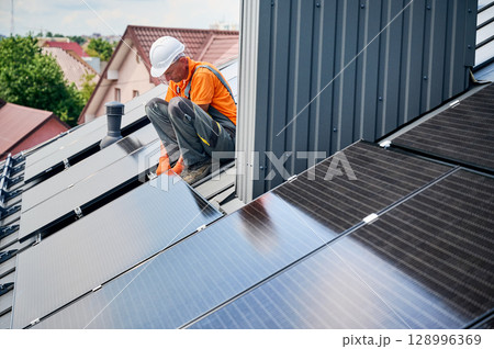 Worker building solar panel system on metal rooftop of house. Engineer installing photovoltaic solar module outdoors. Alternative, green and renewable energy generation concept. Worker building solar panel system on metal rooftop of house. Engineer installing photovoltaic solar module outdoors. Alternative, green and renewable energy generation concept. 128996369