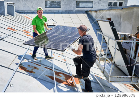 Workers building solar panel system on metal rooftop of house with assistance of crane lift. Two men installers carrying photovoltaic solar module outdoors. Renewable energy generation concept. 128996379