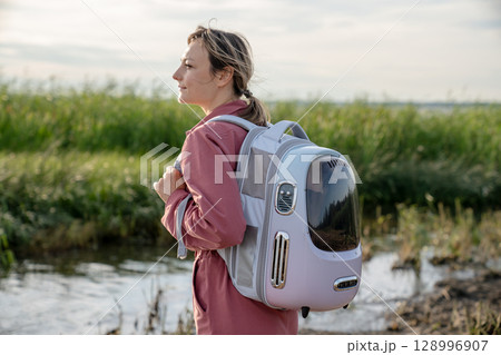 Carefree woman tourist walking riverside trail carrying cat in backpack. Independent travel with pet Carefree woman tourist walking riverside trail carrying cat in backpack. Independent travel with pet 128996907