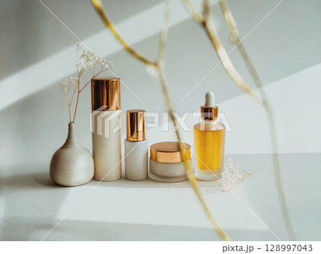 Elegant clean beauty skincare bottles with golden caps and glass dropper in natural sunlight on minimal beige background with dried flowers 128997043
