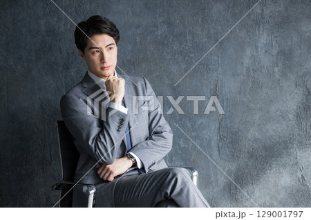 グレーバックで椅子に座るスーツのビジネスマン上半身 129001797