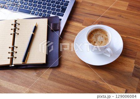 リング式のシステム手帳を広げ、コーヒー(カフェ・ラテ)を飲みながらパソコンでデスクワーク中のイメージ リング式のシステム手帳を広げ、コーヒー(カフェ・ラテ)を飲みながらパソコンでデスクワーク中のイメージ 129006084