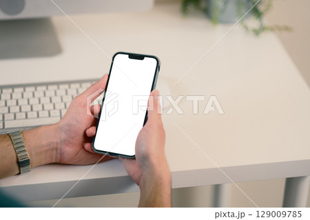 Close up of hands holding smartphone with blank white screen at a modern desk Close up of hands holding smartphone with blank white screen at a modern desk 129009785