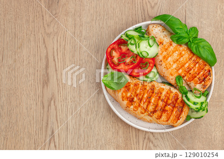 Grilled chicken breast with tomato cucumber salad and basil on wooden background. top view. copy space Grilled chicken breast with tomato cucumber salad and basil on wooden background. top view. copy space 129010254