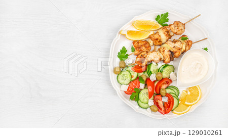 Chicken skewers served with fresh vegetable salad, lemon slices, and creamy sauce on a white plate over a bright wooden background. top view. copy space Chicken skewers served with fresh vegetable salad, lemon slices, and creamy sauce on a white plate over a bright wooden background. top view. copy space 129010261