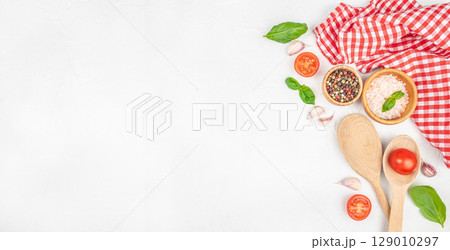 Top view of cooking ingredients with tomato, herbs, spices, and wooden spoons on white background with red checkered cloth 129010297
