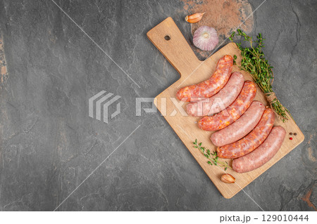 Raw pork sausages with garlic and herbs on a wooden board over a dark stone background. copy space, top view Raw pork sausages with garlic and herbs on a wooden board over a dark stone background. copy space, top view 129010444