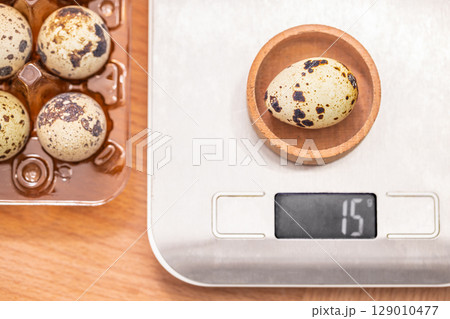 A speckled quail egg is weighed on a digital kitchen scale, displaying a weight of 15 grams beside a plastic egg tray on a wooden surface A speckled quail egg is weighed on a digital kitchen scale, displaying a weight of 15 grams beside a plastic egg tray on a wooden surface 129010477