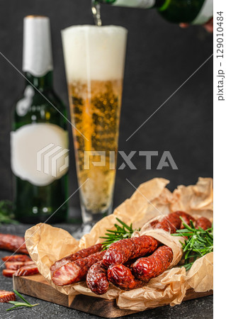 A tall glass of beer is being poured beside a rustic wooden board topped with smoked sausages and sprigs of fresh rosemary, creating a classic pairing of drink and charcuterie 129010484