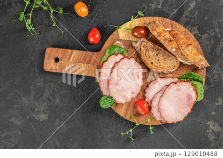 Rustic charcuterie board with sliced ham, whole grain bread, and fresh tomatoes on a dark textured background. top view. copy space 129010488
