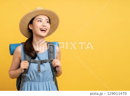 Joyful Asian Backpacker in Straw Hat Gazes Up with Anticipation on Yellow. 129011470