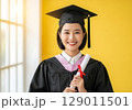 Radiant Graduate in Cap and Gown Holding Diploma Against a Sunlit Yellow Wall. 129011501