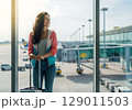 Radiant Traveler with Luggage Gazes Hopefully from Sunlit Airport Window. 129011503