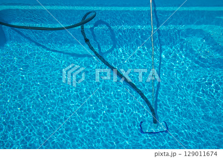 Blue hose and telescopic pole connected to a manual pool vacuum cleaner removing debris from the pool floor during outdoor summer maintenance. 129011674