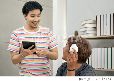 Two young men discussing real estate investment at home office, one smiling while holding tablet and other holding small house model, surrounded by bookshelves in modern indoor 129013473