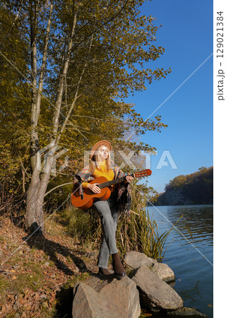 Portrait of smiling caucasian artist plays wooden acoustic guitar in autumnal park. Young songwriter plays string musical instrument hobby outside in nature fall time. Audio music healing 129021384