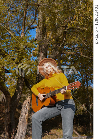Portrait of smiling caucasian artist plays wooden acoustic guitar in autumnal park. Young songwriter plays string musical instrument hobby outside in nature fall time. Audio music healing 129021401