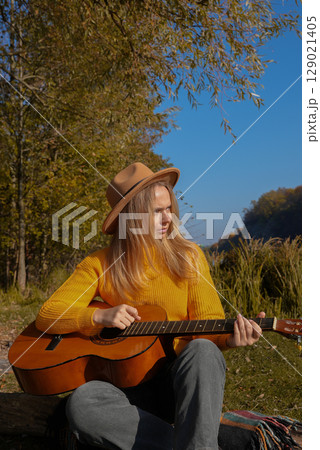 Portrait of smiling caucasian artist plays wooden acoustic guitar in autumnal park. Young songwriter plays string musical instrument hobby outside in nature fall time. Audio music healing Portrait of smiling caucasian artist plays wooden acoustic guitar in autumnal park. Young songwriter plays string musical instrument hobby outside in nature fall time. Audio music healing 129021405