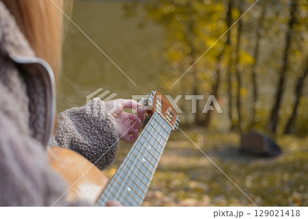 Close up of hands playing acoustic guitar and singing relaxing enjoying outdoors on sunny autumn day. Hippie woman in boho style learning and practicing playing musical instrument. Fall leaves 129021418