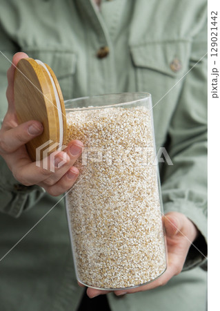 Unrecognizable woman holding refillable glass jar with raw fine ground barley porridge. Refill product format eco-friendly living. Filling reusable container Whole spelt grain species. High protein 129021442