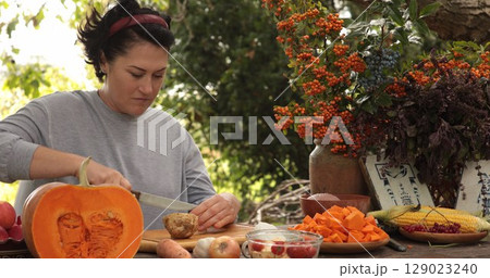 Woman is focused on preparing Thanksgiving meal in outdoor kitchen, chopping fresh vegetables and fruits. Surroundings include autumn foliage and harvest decorations, enhancing festive mood. 129023240