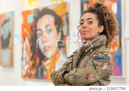 Artist is posing with folded arms in an art gallery, with colorful abstract paintings in the background 129027798
