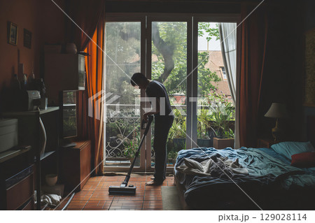 Young man is vacuuming the floor of his bedroom, enjoying the natural light streaming in from the balcony 129028114