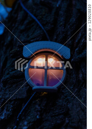 Illuminated decorative light fixture attached to a tree trunk, showcasing a warm glow with a cylindrical transparent shade designed for outdoor ambiance enhancement. Illuminated decorative light fixture attached to a tree trunk, showcasing a warm glow with a cylindrical transparent shade designed for outdoor ambiance enhancement. 129028810