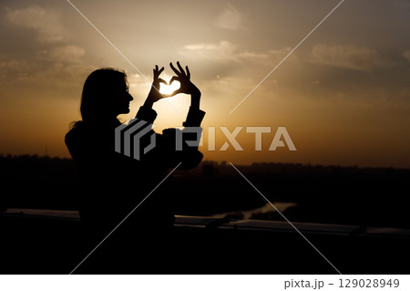Silhouette of a Woman Making a Heart Gesture Against a Golden Sunset Sky with Cloud Patterns Beautifully Framing the Scene, Evoking Love and Serenity 129028949