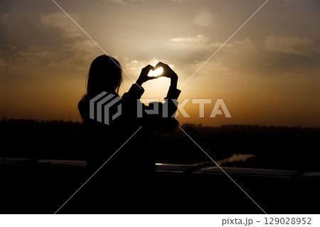 Silhouette of a Person Forming a Heart Shape with Their Hands Against a Beautiful Sunset Over the City Skyline 129028952