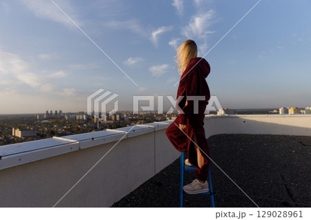 A Woman on a Rooftop at Sunset, Capturing the Beauty of the Urban Skyline Beneath the Warm Glow of the Evening Light with a Sense of Adventure and Freedom in Her Attire A Woman on a Rooftop at Sunset, Capturing the Beauty of the Urban Skyline Beneath the Warm Glow of the Evening Light with a Sense of Adventure and Freedom in Her Attire 129028961