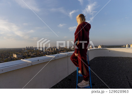 A Young Woman in a Cozy Outfit Enjoys the Scenic Rooftop Views of the City at Sunset, Embracing the Calm before the Night in a Beautiful Urban Landscape. 129028963