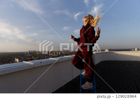 A Young Woman Standing on a Chair on a Rooftop Enjoys the Breeze in a Cozy Outfit While Overlooking an Urban Landscape Under a Beautifully Cloudy Sky 129028964