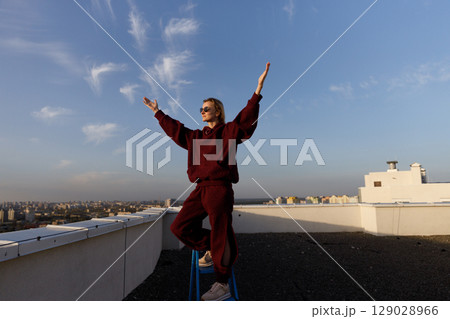 A Confident Young Woman Embraces the Skyline from a Rooftop, Dressed in a Comfy Stylish Outfit While Hailing the Endless Possibilities of the Urban Cityscape 129028966