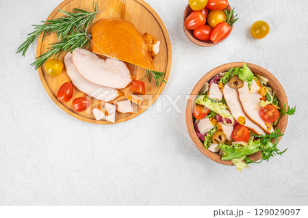 Fresh salad with sliced smoked turkey breast, cherry tomatoes, and olives served alongside a wooden board of turkey and herbs. top view. copy space. 129029097