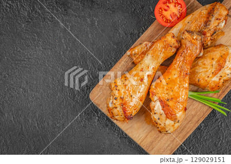 Juicy roasted chicken drumsticks served on a wooden board with tomato and green onion garnish on a dark background. top view. copy space 129029151