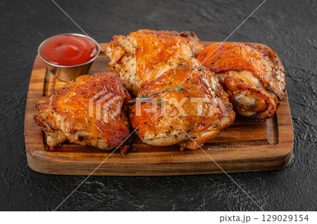 Juicy roasted chicken thighs with crispy skin served on a wooden board with ketchup and herbs on a dark textured background 129029154