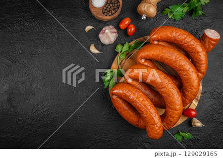 smoked sausages on a wooden board with herbs, garlic, pepper, and tomatoes on a dark textured background. top view. copy space 129029165