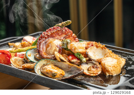 A hot iron skillet sizzles with a vibrant seafood medley including octopus, mussels, clams, shrimp, scallops, tomatoes, and asparagus in a rich savory sauce. A hot iron skillet sizzles with a vibrant seafood medley including octopus, mussels, clams, shrimp, scallops, tomatoes, and asparagus in a rich savory sauce. 129029168