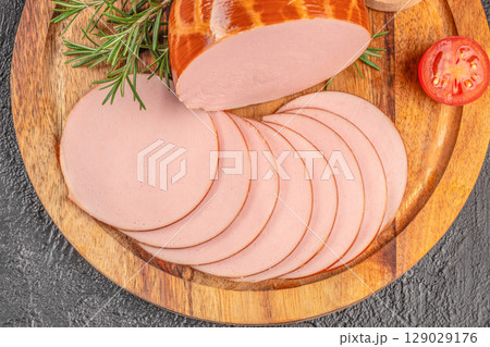 Sliced bologna sausage arranged on a wooden board with rosemary and tomato on a dark background 129029176