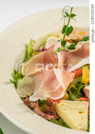 Thin slices of prosciutto rest on a bed of fresh mixed greens with citrus segments and soft cheese, creating a vibrant and savory salad presentation Thin slices of prosciutto rest on a bed of fresh mixed greens with citrus segments and soft cheese, creating a vibrant and savory salad presentation 129029299